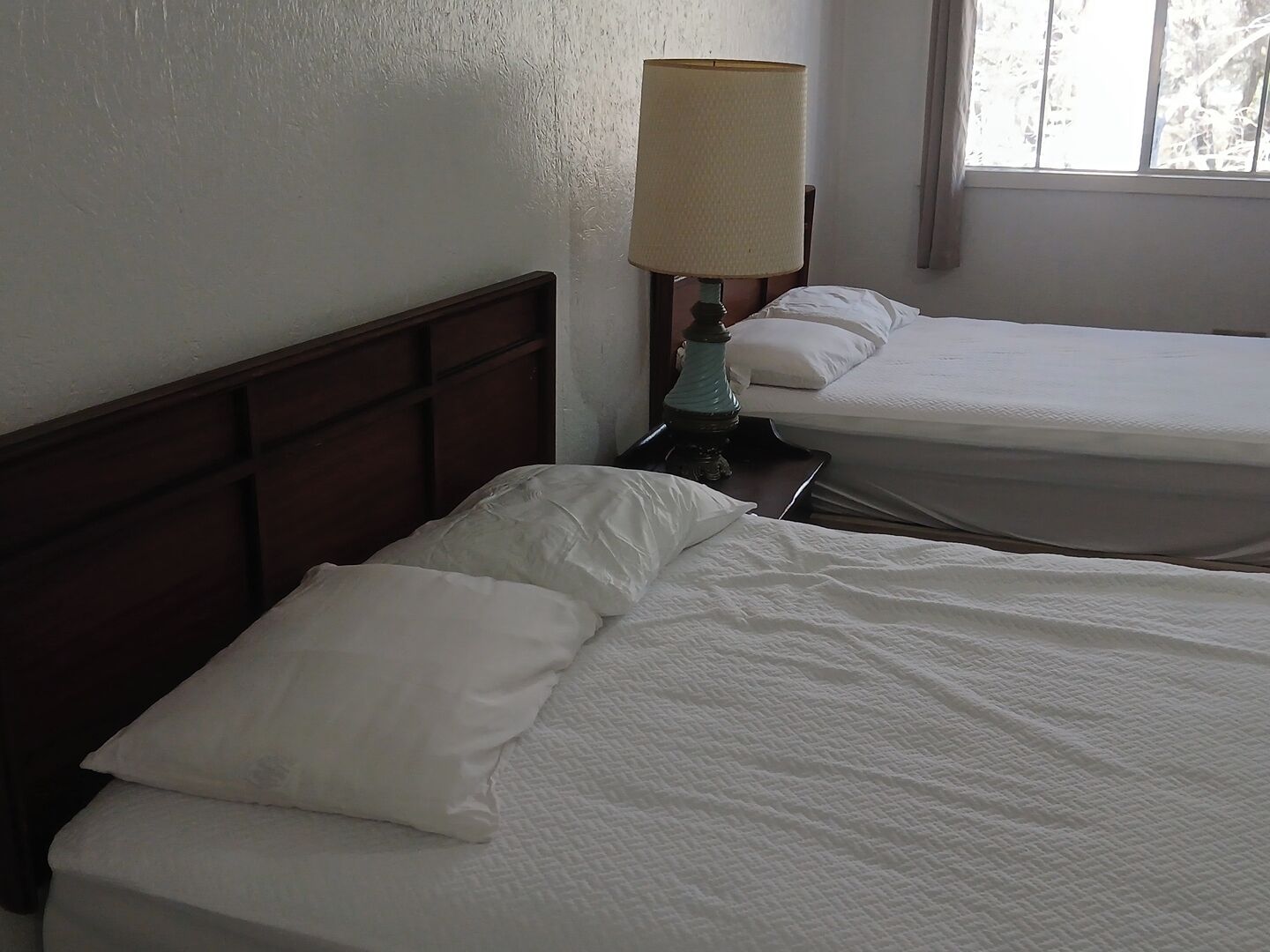 A hotel room with two beds, a nightstand with a lamp, and a window with blinds.