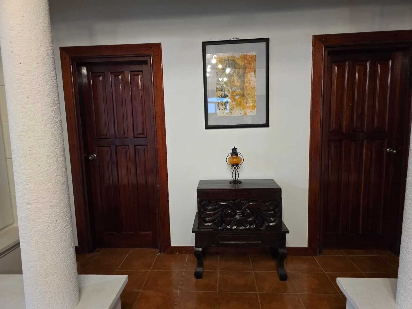 A hallway with two wooden doors, a framed abstract painting, and a small table with a lamp.