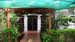 Een overdekt buitengebied met een houten pergola, een pad naar een deur en weelderige begroeiing.