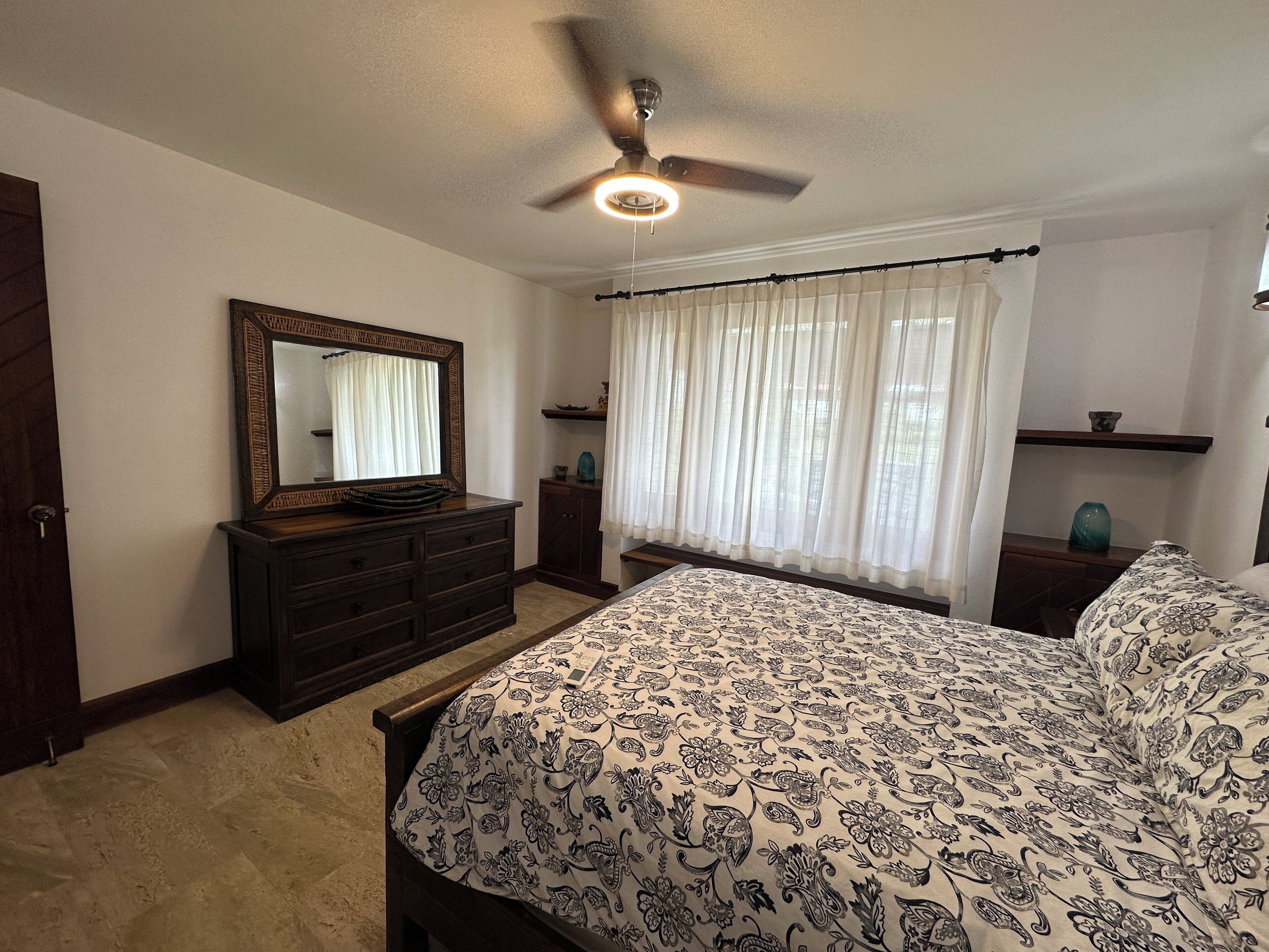 A bedroom with a bed, dresser, mirror, ceiling fan, and window with sheer curtains.