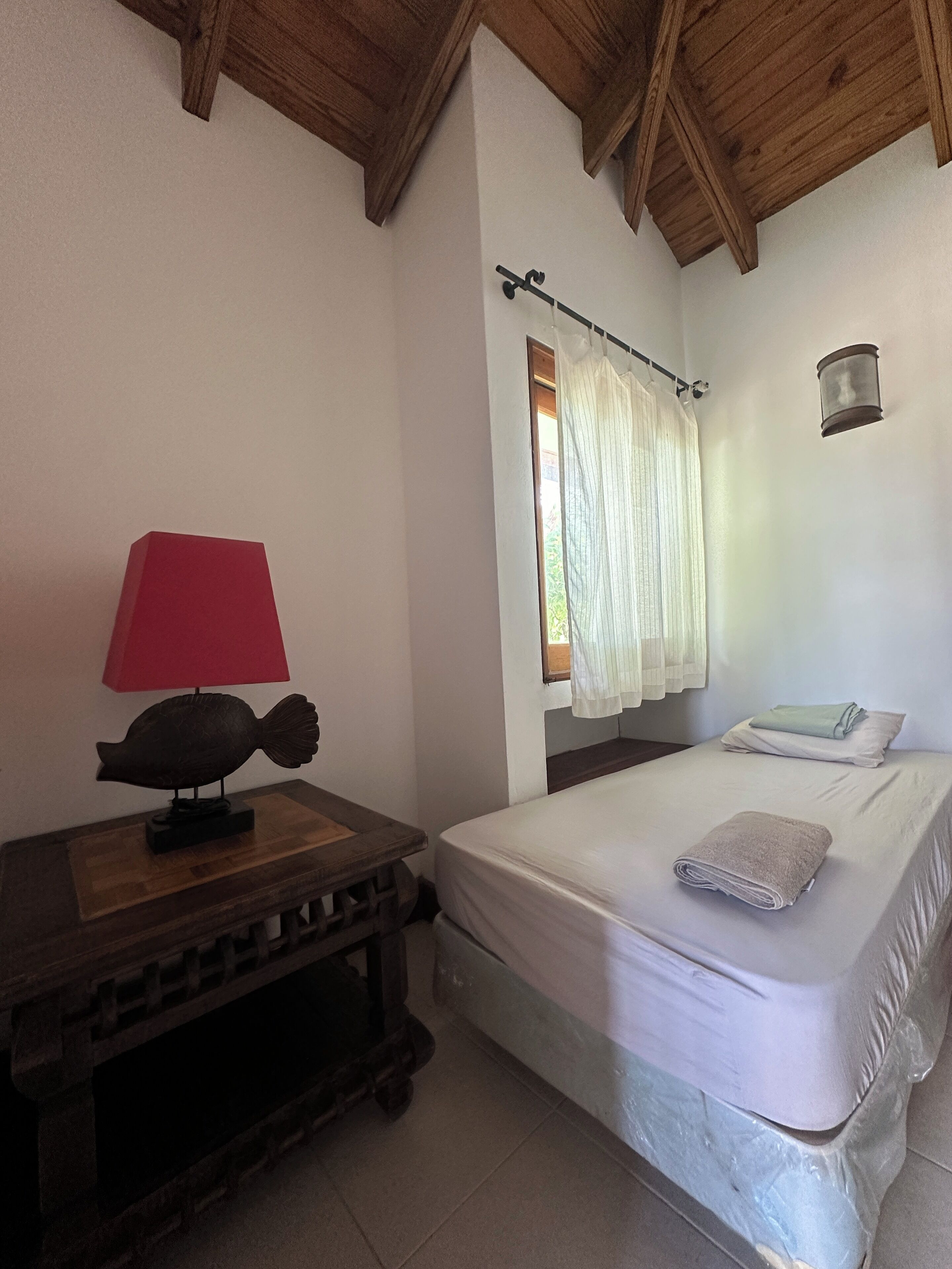A bedroom with a wooden headboard, a bed with a white mattress and a folded towel, a bedside table with a lamp, and a window with curtains.