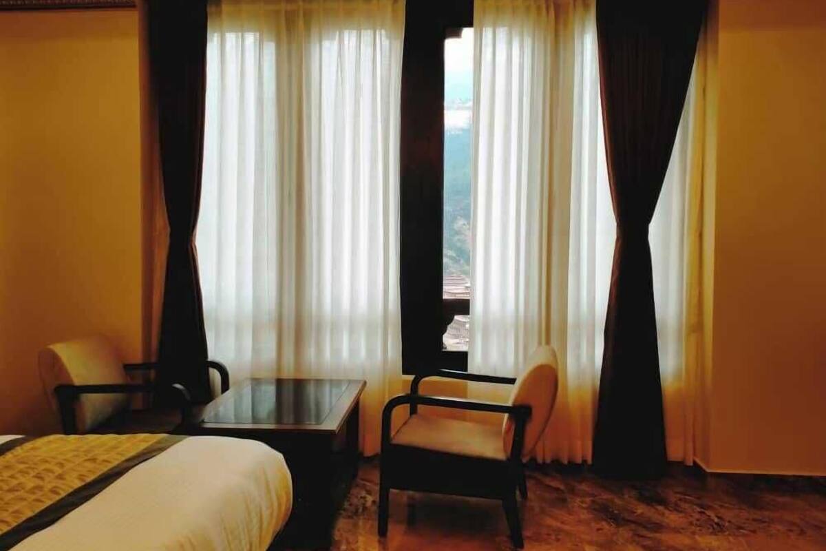 Une chambre d’hôtel avec un lit, deux chaises, une petite table et une vue sur un paysage montagneux, visible à travers de fines rideaux.