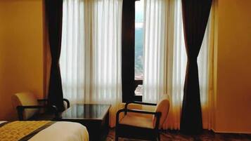 Habitación de hotel con una cama, dos sillas, una mesita y vistas a un paisaje montañoso a través de cortinas translúcidas.