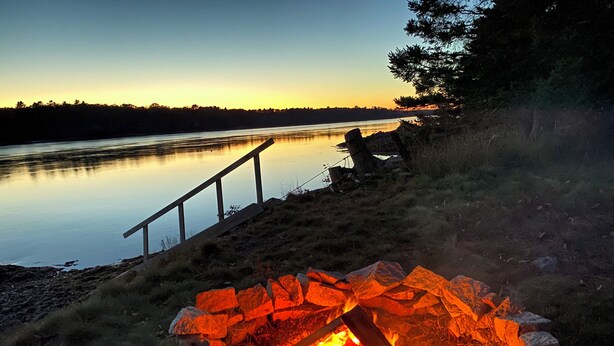 A campfire surrounded by rocks, a wooden railing, and a calm lake at dusk.