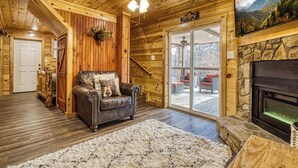 A cozy cabin interior with a fireplace, wooden walls, a leather sofa, and a large window overlooking a deck. - Hidden Haven, Hot Tub, FREE Attraction tickets each paid day, Game Room, WIFI!!! (Sevierville)