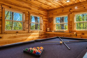 Una mesa de billar en una sala de madera con ventanas grandes y vistas a árboles.