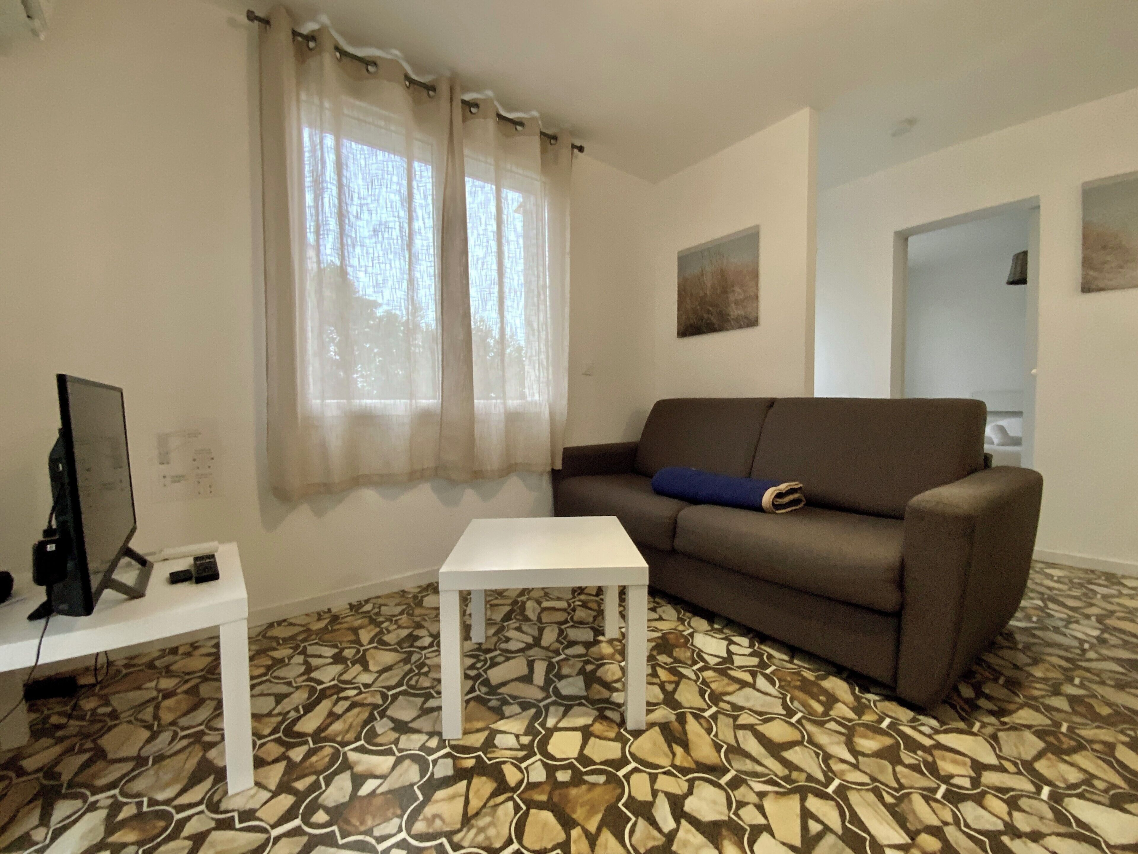 Ein Wohnzimmer mit einem gemusterten Boden, einem braunen Sofa, einem weißen Tisch und einem Fernseher auf einem Lowboard.