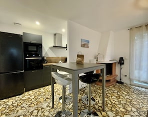 Cocina compacta con refrigerador, horno y microondas. Hay una mesita con sillas, una cafetera y un bolso sobre la encimera. El piso tiene un diseño de baldosas con motivos.
