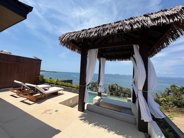 A sunlit outdoor lounge area with a view of the sea, a thatched-roof structure, and a pool.