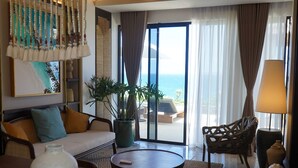 Habitación de hotel con sofá, una mesa redonda, una silla, planta en maceta y vista al océano a través de amplias ventanas.
