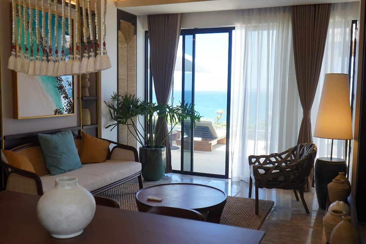 A hotel room with a sofa, a round table, a chair, a potted plant, and a view of the ocean through large windows.