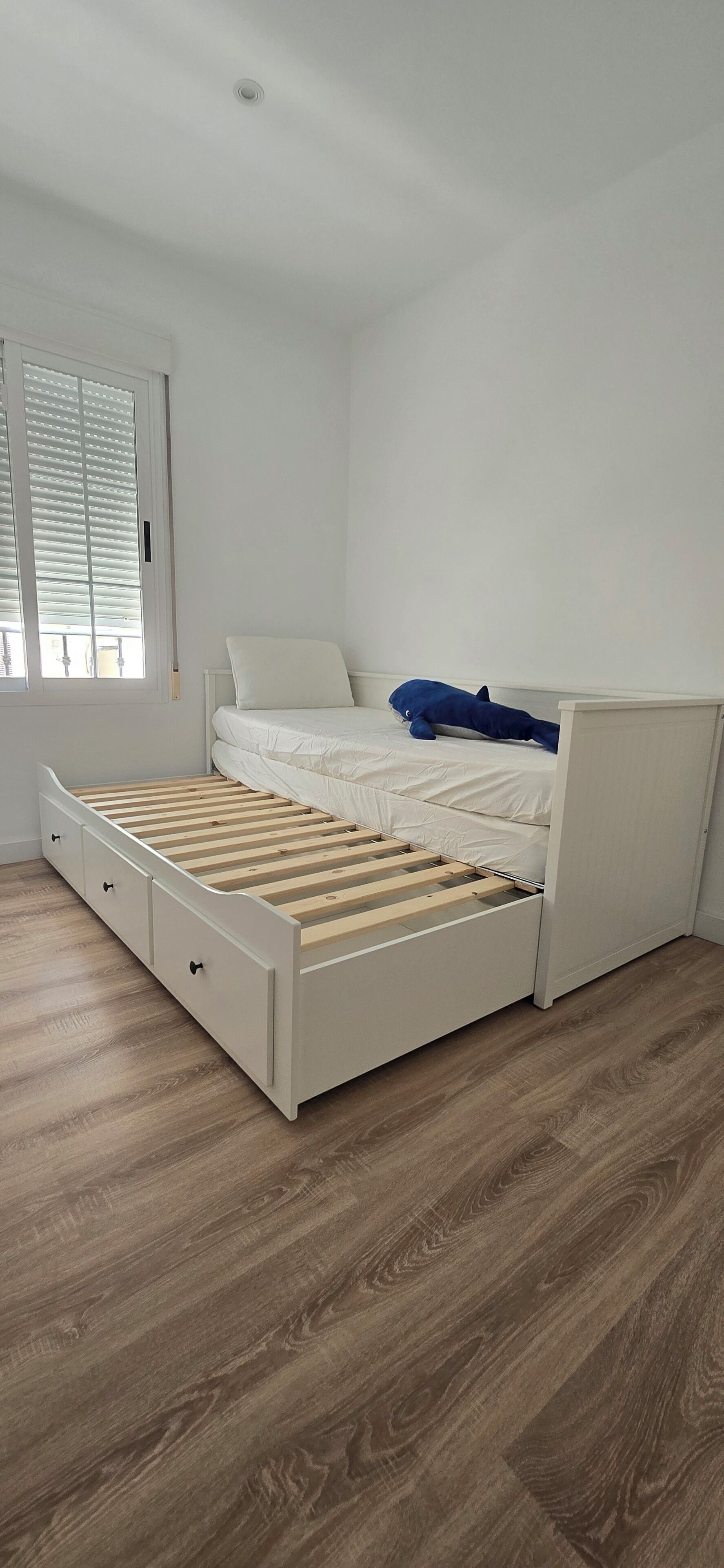 Ein weißes Bett mit Holzrahmen, eine weiße Matratze und ein blaues Kopfkissen. Ein Fenster mit Jalousien und eine weiße Wand sind zu sehen.