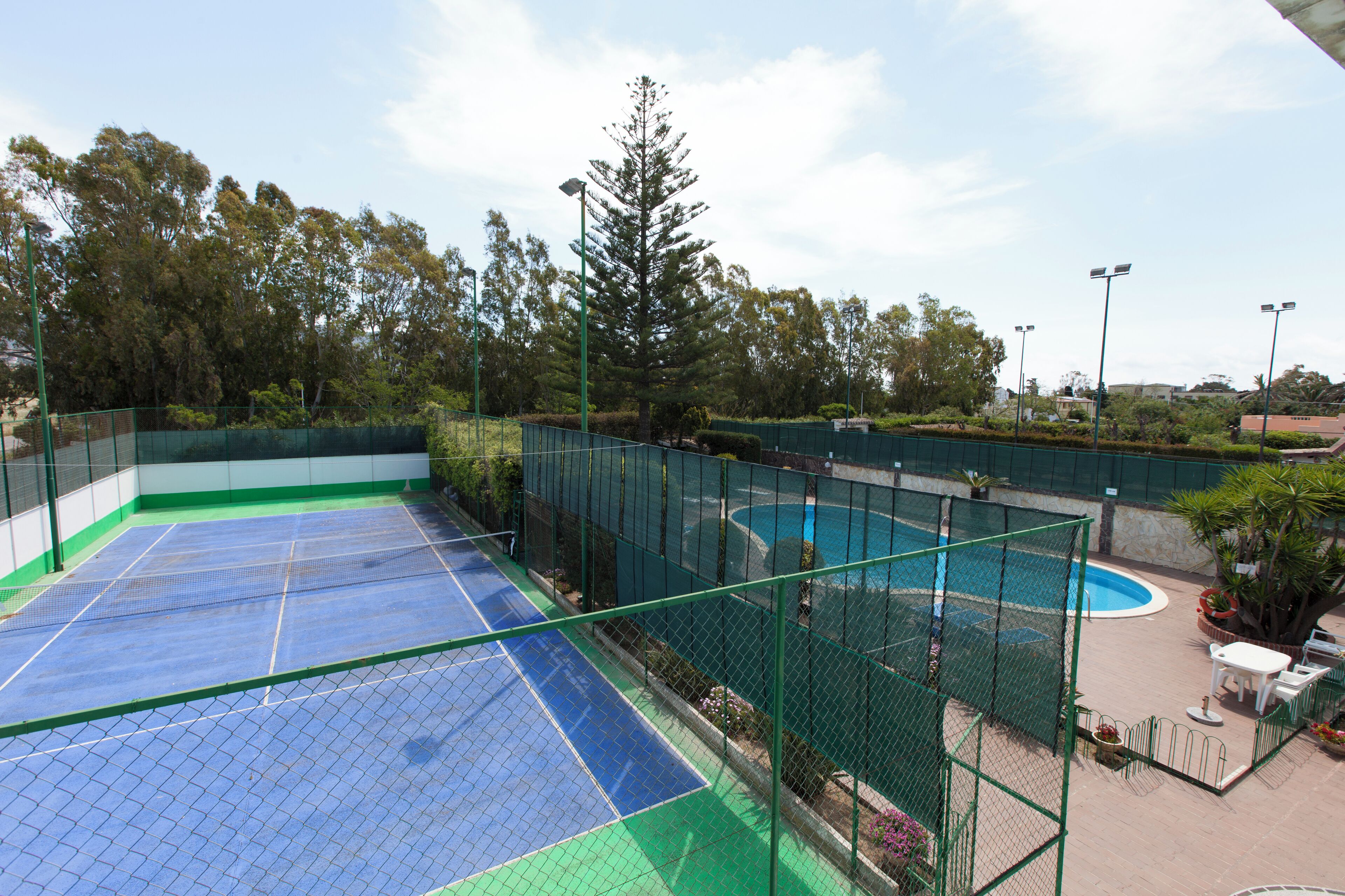 Una cancha de tenis al aire libre con piscina, rodeada por una cerca verde y árboles.