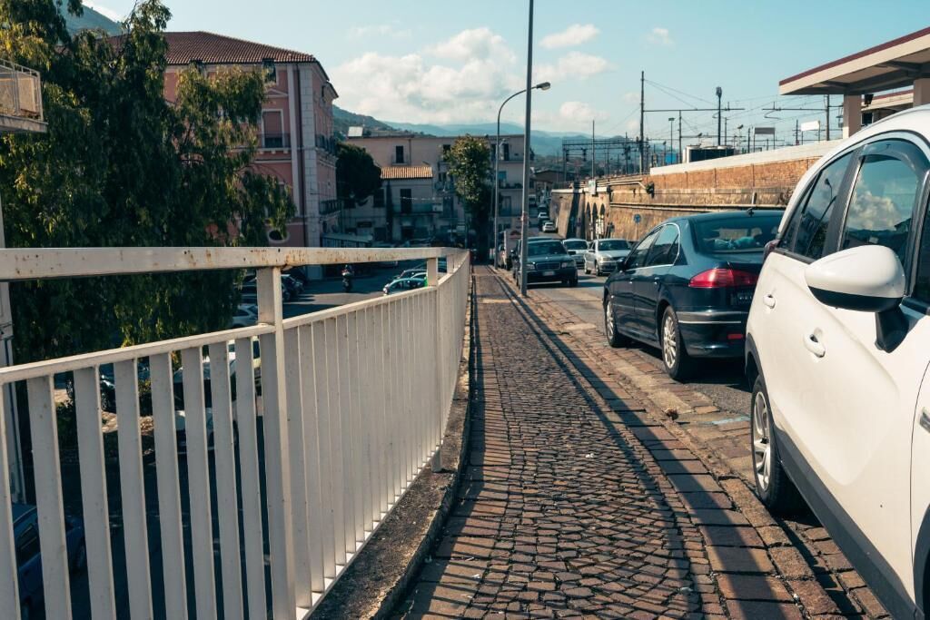A cobblestone street with parked cars and a white fence on one side.