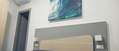 A hotel room with a bed, a wall-mounted air conditioner, a painting of a wave, and a door.