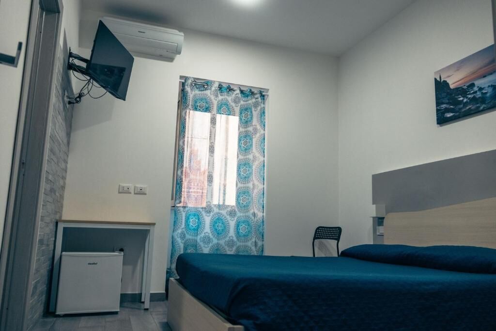 Un dormitorio con cama, escritorio, silla, televisor montado en la pared y ventana con cortinas estampadas.