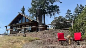 A log cabin with a large chimney, a deck with red chairs, and a forested backdrop. - Incredible Views - Orr Springs Rendezvous! (Ukiah)