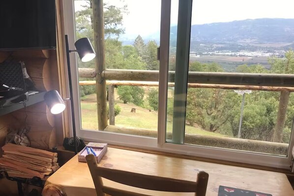 A room with a wooden desk, a chair, and a large window offering a view of a forest and distant buildings.