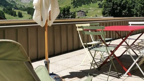 Ein Balkon mit Blick auf grüne Berge, einem weißen Sonnenschirm und einem roten Tisch mit Stühlen.