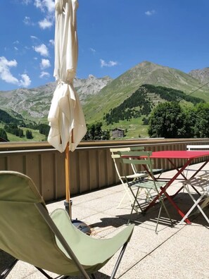 En balkon med udsigt til grønne bjerge, en hvid parasol samt et rødt bord og stole.