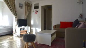 Ein Wohnzimmer mit Bett, Fernseher auf einem Schrank, einem weißen Couchtisch, einem roten Kissen und einer Couch.