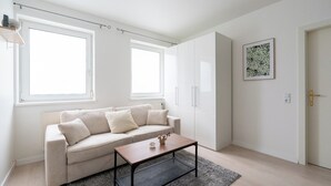 Un salon moderne avec un canapé beige, une table basse en bois et un tapis gris. Il y a deux fenêtres qui laissent entrer la lumière naturelle, et une porte sur le côté droit.