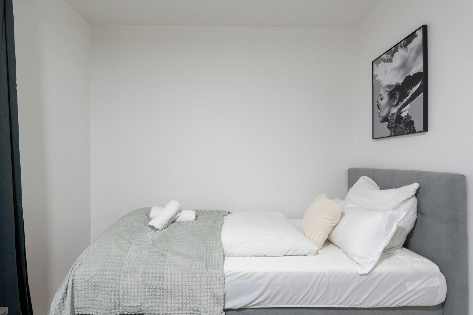 Una cama bien hecha con sábanas blancas, un cabecero gris y una manta gris, con un solo almohadón blanco.