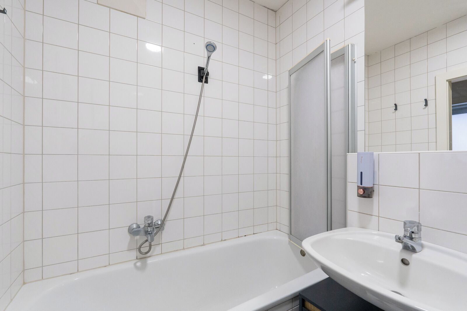 Et badeværelse med hvide flisevægge, badekar, bruser og et spejl over håndvasken.