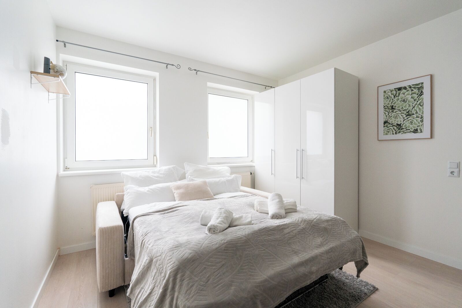 Un dormitorio moderno con cama, dos ventanas, un radiador, un armario y una obra de arte floral enmarcada en la pared.