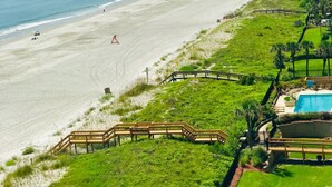 Ein Strand mit einer hölzernen Seebrücke, die ins Meer führt, einem Rasenbereich mit einem Pool und einem klaren Himmel.