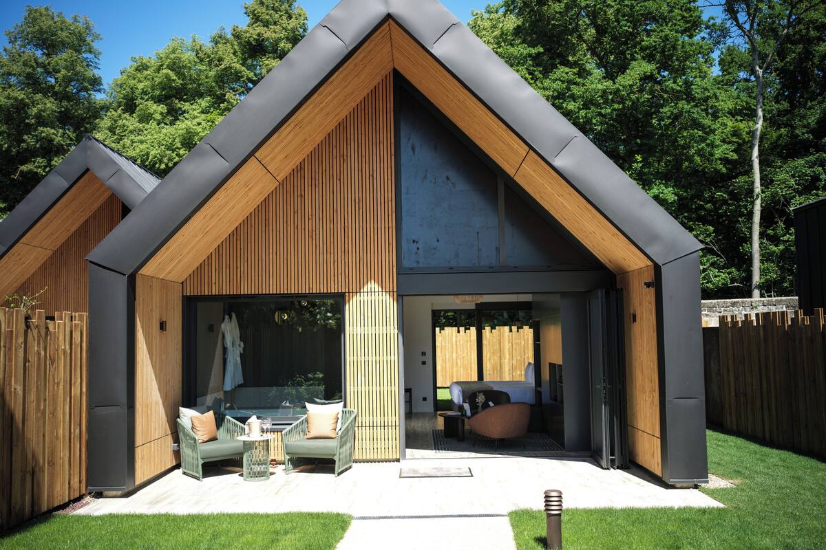 A modern house with a sloped roof, wooden exterior, and a large glass door leading to a living area with a sofa and a dining table.
