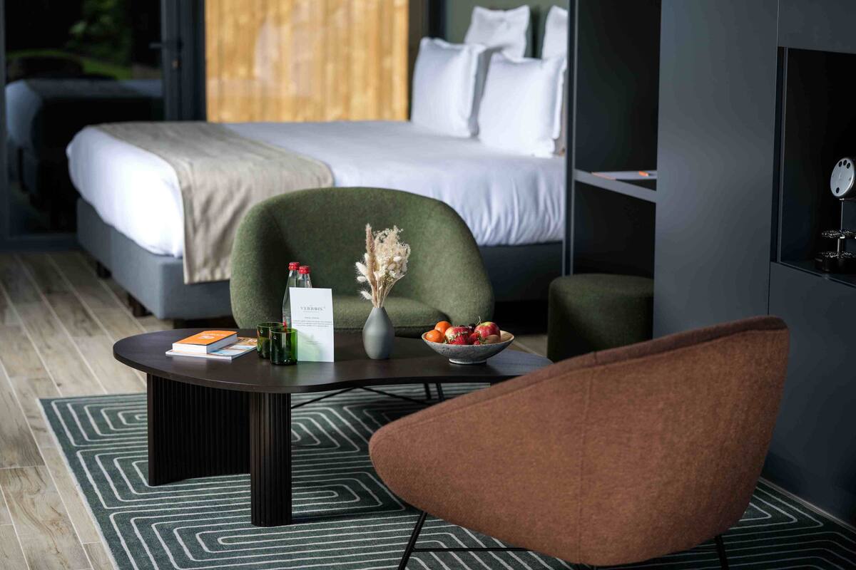 A modern hotel room with a bed, a green armchair, a coffee table with a vase and fruit bowl, and a small table with books and glasses.