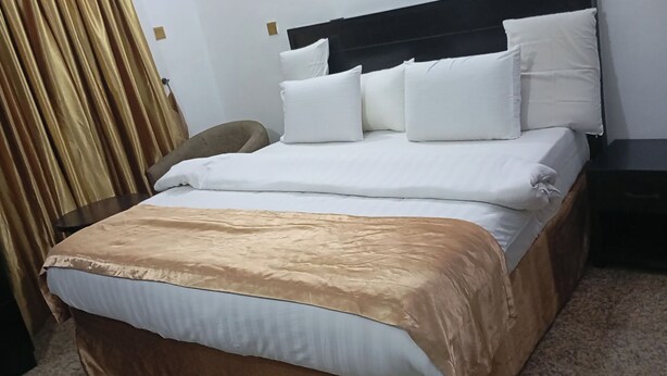 Ein ordentlich bezogenes Bett mit weißen Laken und einer braunen Tagesdecke, ein dunkles Kopfbrett sowie ein Nachttisch mit einer Lampe.