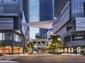 Una calle urbana moderna con edificios altos, un puente peatonal y tiendas, incluyendo una tienda Cole Haan.