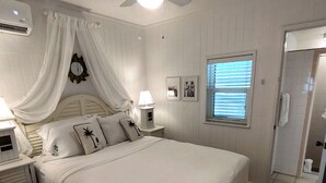 Une chambre à coucher comprenant un lit, un ventilateur de plafond, une lampe de chevet, une fenêtre avec des stores et une porte.