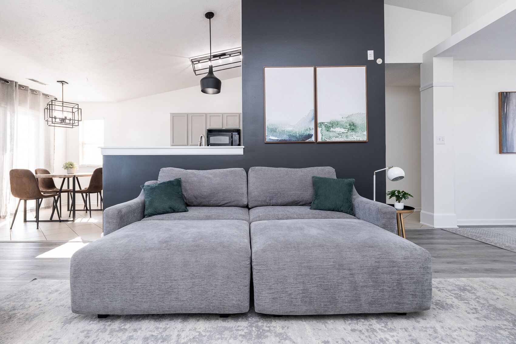 Una sala moderna con un sofá seccional gris, un comedor con mesa y sillas, y una cocina al fondo.