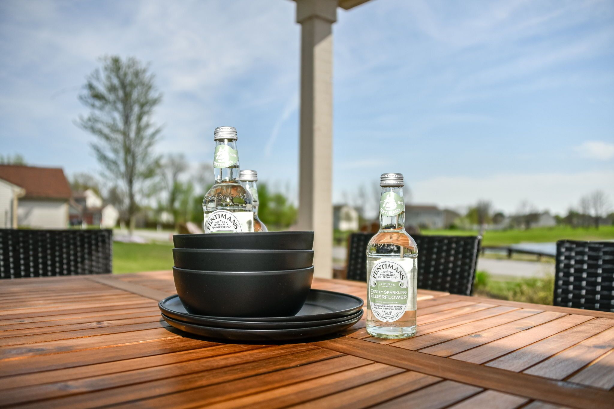 Taula de fusta amb tres ampolles de Fentimans Cidere Sparkling Elderflower sobre posavasos negres, situada a l'exterior amb un cel clar i vegetació al fons.
