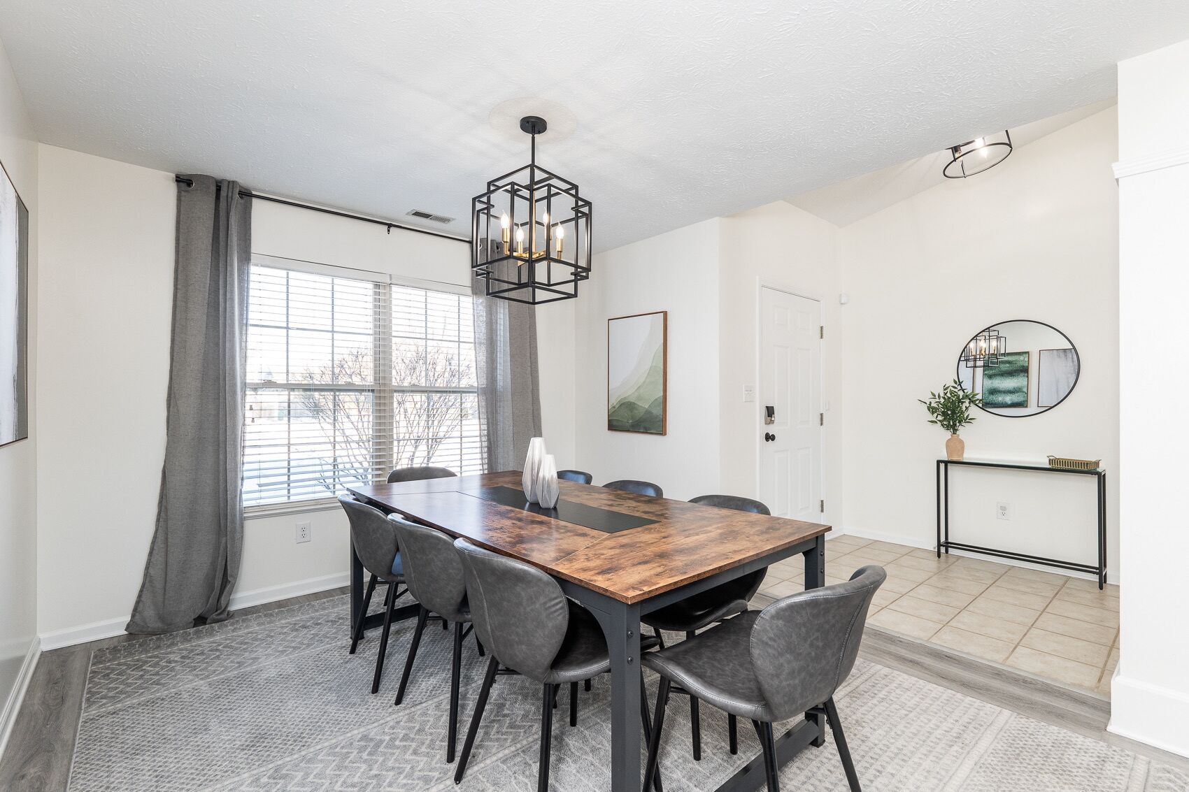 A dining area with a wooden table, six chairs, a hanging light fixture, a white door, and a console table with a mirror and plant.