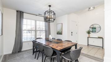 A dining area with a wooden table, six chairs, a hanging light fixture, a white door, and a console table with a mirror and plant.