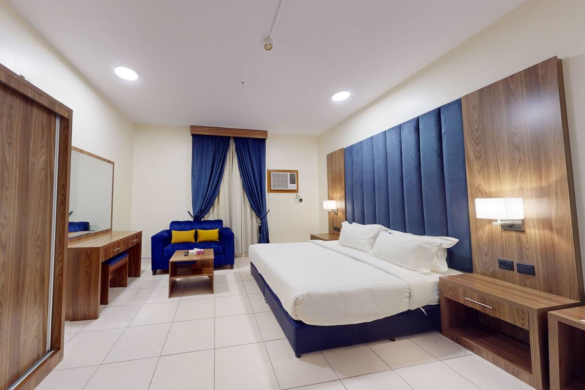 A hotel room with a large bed, a desk with a mirror, a blue sofa, and a wooden wardrobe.