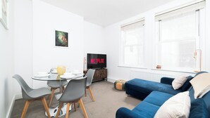 A modern living room with a blue sofa, a glass coffee table, and a dining area with chairs. A television displaying 'NETFLIX' is visible in the background. - 
Stylish Apartment Close to Fulham Palace (London)