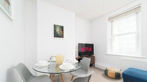 A modern living room with a glass dining table, grey chairs, a blue sofa, a television, and a framed picture on the wall. - 
Stylish Apartment Close to Fulham Palace (London)