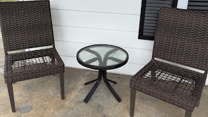Two wicker chairs and a small round table with a glass top, positioned on a concrete patio against a white house wall with black shutters. - Downtown Convenience near Lake Norman (Mooresville)