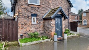 A brick house with a dark blue door, a small window, and a garden area with plants and a pathway. - Cozy Trafford Cottage Just 14 Minutes from Airport (Partington)