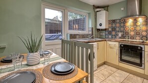 A compact kitchen with a dining area, featuring a tiled backsplash, a built-in oven, and a window providing natural light. - Cozy Trafford Cottage Just 14 Minutes from Airport (Partington)