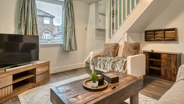 A living room with a TV on a wooden stand, a sofa with a fur throw, a coffee table with a plant, and a staircase with a wooden railing. - Cozy Trafford Cottage Just 14 Minutes from Airport (Partington)