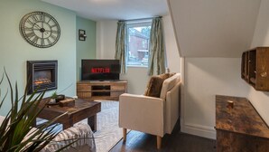 A living room with a fireplace, a television, a clock, and a window with curtains. - Cozy Trafford Cottage Just 14 Minutes from Airport (Partington)
