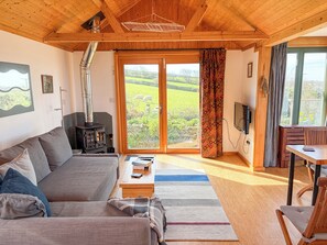 A cozy living room with a sofa, a wooden table, a TV, and a large window with a view of a green landscape.