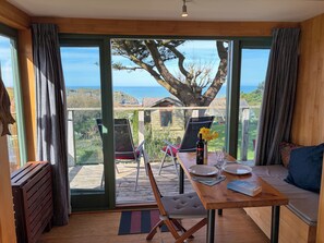 A room with a wooden floor, a dining table with chairs, a sofa, and a view of the ocean through large windows. - Quiet chalet with beautiful rural and sea views near St Nectan's waterfall. (Trethevy)