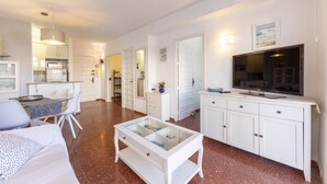Een moderne woonkamer met een salontafel met glazen blad, witte banken, een eethoek met tafel en stoelen, een keuken met witte kasten, en een televisie op een witte kast.